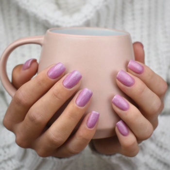 Zbliżenie dłoni trzymających kubek, z liliowym lakierem hybrydowym na paznokciach, z efektem brokatowego pyłku.