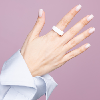 Biały, szeroki pierścionek na palcu serdecznym kobiecej dłoni z delikatnym manicure, na fioletowym tle.