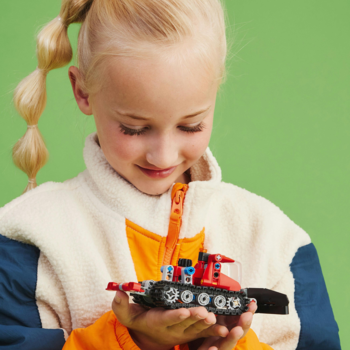 Dziewczynka w kurtce trzyma czerwony LEGO Technic Ratrak 42148. Zbliżenie na dłonie i model na zielonym tle.