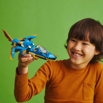 Dziecko trzyma w ręku niebiesko-złoty LEGO Odrzutowiec ponaddźwiękowy Jaya EVO, uśmiecha się na zielonym tle.