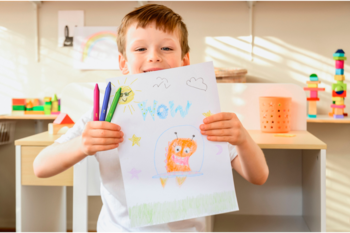 Uśmiechnięty chłopiec z rysunkiem kosmity i zestawem kredek świecowych, pokazujący radość tworzenia.