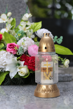 Znicz nagrobny ze złotym lampionem i białym wkładem nr 3 (55h palenia) na nagrobku, obok kwiaty.