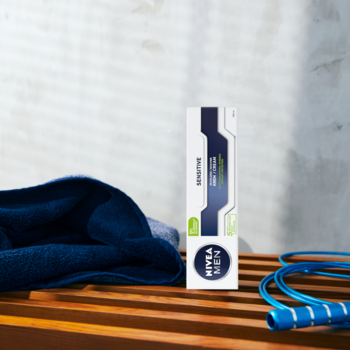 NIVEA MEN Sensitive krem do golenia w biało-niebieskim opakowaniu, stoi na drewnianej ławce obok ręcznika i skakanki.