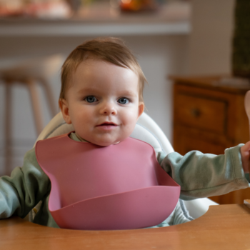 Uśmiechnięte niemowlę z niebieskimi oczami, w różowym silikonowym śliniaku, siedzi gotowe na BoboVita kaszkę.