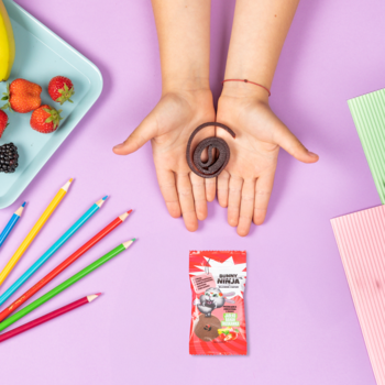 Dziecięce ręce prezentują spiralną, suszoną przekąskę owocową. Obok czerwona paczka jabłko, banan, truskawka.