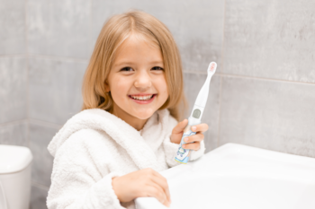 Uśmiechnięta blond dziewczynka w szlafroku prezentuje soniczną szczoteczkę Vitammy Smile w łazience.