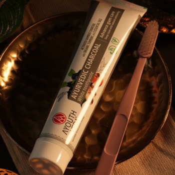 Wybielająca pasta AYOUTH Ayurvedic Charcoal z aktywnym węglem w tubce i różowa szczoteczka na złotej tacy, z góry.