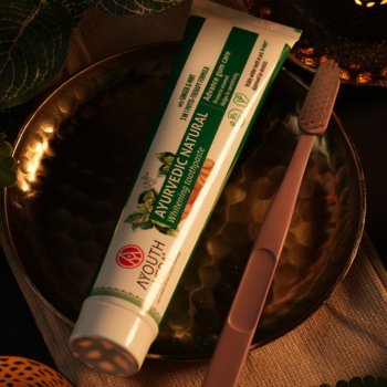 Pasta AYOUTH Ayurvedic Natural Whitening, imbir i mięta, tubka obok różowej szczoteczki na złotej tacy.