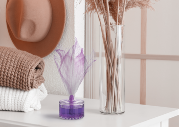 Lawendowy dyfuzor zapachowy z fioletowym kwiatem na białym blacie. W tle stos koców i wazon z trawą. Widok z przodu.