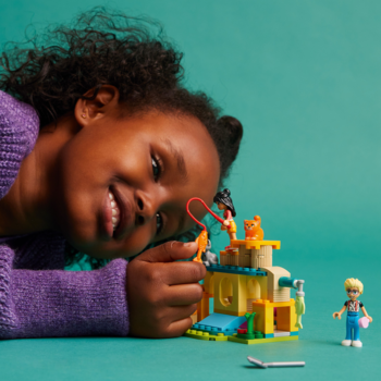 Uśmiechnięta dziewczynka buduje zestaw LEGO Friends Przygoda na kocim placu zabaw. Widok z boku, turkusowe tło.