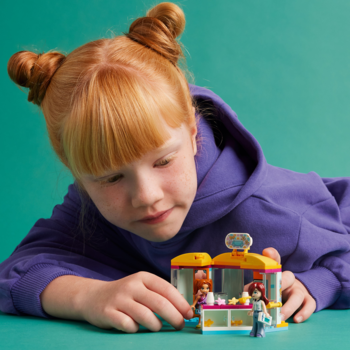 Ruda dziewczynka w fioletowej bluzie bawi się Lego Friends Mały sklep z akcesoriami, budując scenki z laleczkami.
