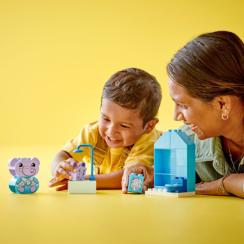 Uśmiechnięte dziecko z dorosłym budują Lego Duplo Codzienne Czynności. Widoczne słoniki, wanna, prysznic na żółtym tle.