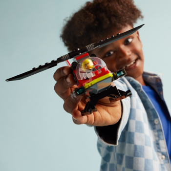 Uśmiechnięty chłopiec trzyma czerwony Lego strażacki helikopter ratunkowy z figurką pilota, z przodu.