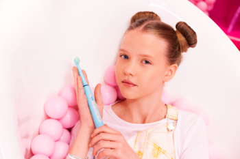 Młoda dziewczynka w białej wannie z różowymi kulkami trzyma jasnoniebieską szczoteczkę soniczną VITAMMY Splash.
