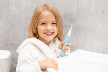 Uśmiechnięta dziewczynka w białym szlafroku trzyma niebieską szczoteczkę soniczną VITAMMY SMILE w łazience.