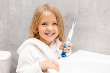 Uśmiechnięta dziewczynka w białym szlafroku trzyma VITAMMY SMILE, dziecięcą szczoteczkę soniczną.