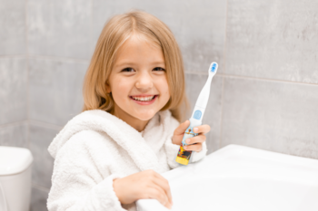 Uśmiechnięta dziewczynka w białym szlafroku, trzyma kolorową soniczną szczoteczkę Vitammy Smile dla dzieci.