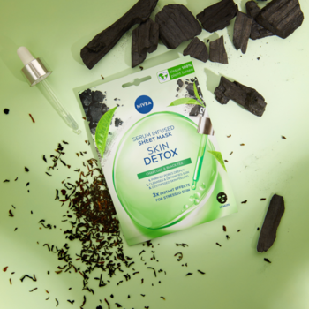 Maska w płachcie Nivea Skin Detox z serum, aktywnym węglem i herbatą, na zielonym tle z węglem i liśćmi herbaty.