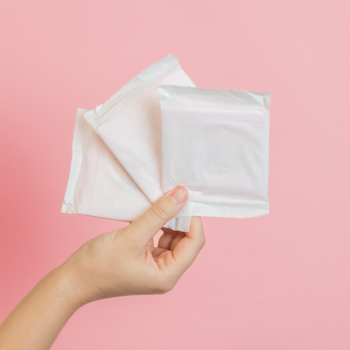 Dłoń prezentuje trzy białe podpaski JONI TEENS, pakowane osobno, na jasnoróżowym tle. Hipoalergiczne, 100% bawełny organicznej.