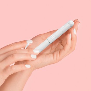 Biały tampon JONI TEENS mini z kartonowym aplikatorem, bawełna organiczna, w dłoniach na różowym tle.