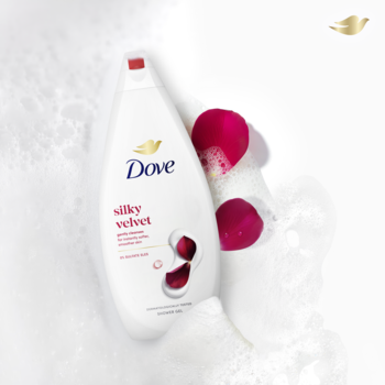 Dove Silky Velvet żel pod prysznic, biała butelka z czerwonymi akcentami, leży na pianie z płatkami róż.