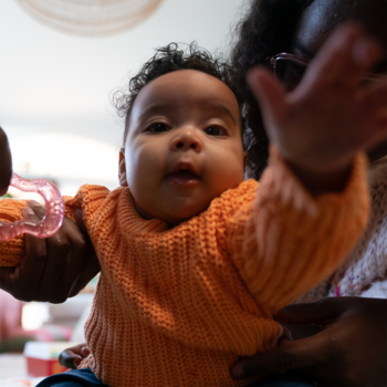Ujęcie bliskie radosnego niemowlęcia w pomarańczowym swetrze z gryzakiem, Bobovita dla dzieci po 6 m-cu.