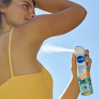 Kobieta aplikuje antyperspirant NIVEA Tropical Sunrise w sprayu, butelka z egzotycznymi owocami. Widoczna mgiełka, 72h ochrony.