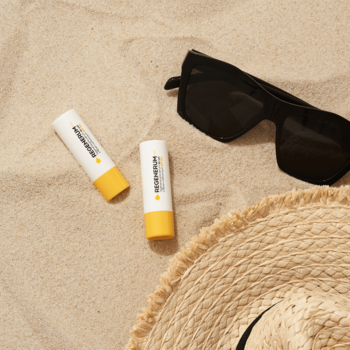 Dwa serum do ust Regenerum SPF 50+ na piasku, widok z góry, obok słomkowy kapelusz i okulary.