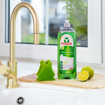 Frosch Limonkowy płyn do naczyń, zielona butelka przy zlewie. Obok zielona gąbka-żabka i świeże limonki.