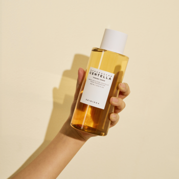 Ręka podnosi przezroczystą butelkę toniku SKIN1004 Madagascar Centella Toning Toner z bursztynowym płynem.