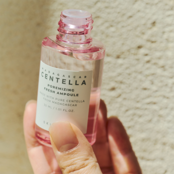 Skin1004 Centella Poremizing Fresh Ampoule, różowe serum w małej szklanej butelce, w dłoni na jasnym tle.