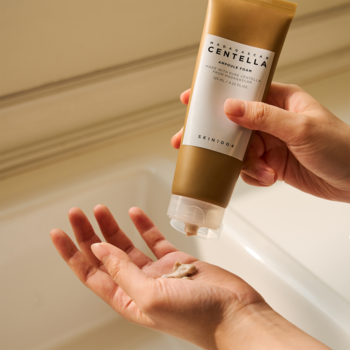 Beżowa tuba Skin1004 Madagascar Centella Ampoule Foam, z której wyciskana jest pianka na dłoń w jasnej łazience.