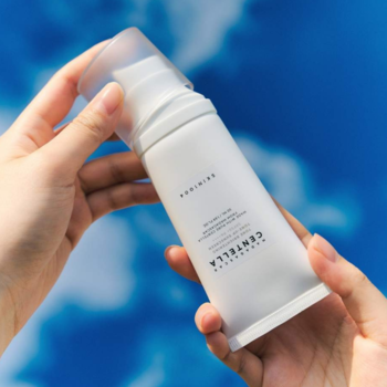 Biała butelka kremu ochronnego SPF 50+ z Centella Asiatica, trzymana w dłoniach na tle błękitnego nieba.