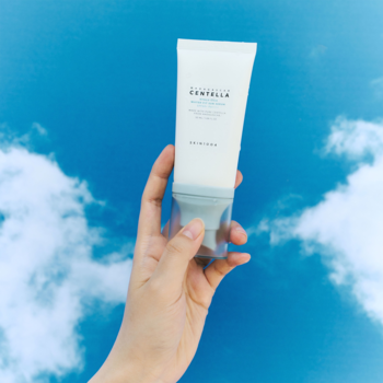 Biała tubka Skin1004 Madagascar Centella Hyalu-Cica Water-Fit Sun Serum SPF50+ w dłoni, na tle niebieskiego nieba z chmurami.