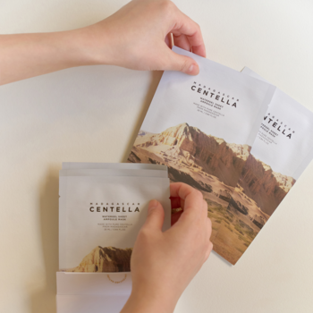 Dłonie układające białe, kojąco-nawilżające maski hydrożelowe z centellą i górskim motywem, widok z góry.