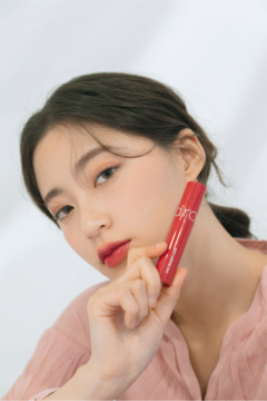 Kobieta prezentuje czerwony błyszczyk Rom&nd Juicy Lasting Tint nr 06 w tubce, blisko twarzy, białe tło.