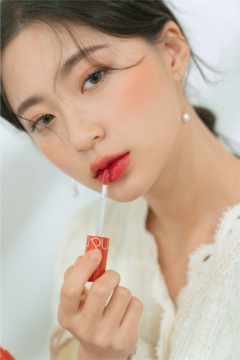 Zbliżenie na kobietę aplikującą błyszczyk Rom&nd Juicy Lasting Tint nr 07 w odcieniu koralowym, z połyskiem.