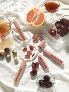 Rom&nd Juicy Lasting Tint nr 25: beżowo-różowe błyszczyki, swatche, owoce i napoje na białej tkaninie.