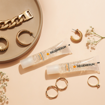Regenerum serum do ust (żel-olejek) w dwóch przezroczystych tubkach, otoczone złotą biżuterią na beżowym tle.