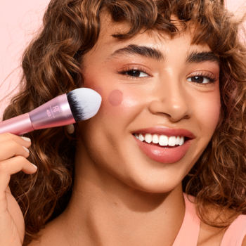 Kobieta nakłada róż na policzek pędzlem Real Techniques Power Pigment Blush z różową rączką i białym włosiem.