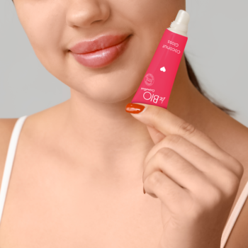 Różowy żelowy olejek do ust Coconut Gloss w tubce, trzymany przez kobietę. Widoczne nawilżone, pełne usta.