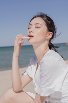 Kobieta na słonecznej plaży aplikuje Rom&nd Zero Matte Lipstick w odcieniu Shell Nude na usta, widać matowe wykończenie.