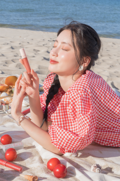 Rom&nd Zero Velvet Tint Burny Nude matowa pomadka, trzymana przez kobietę na plaży, na czerwono-białym kocu.