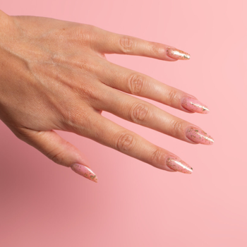 Manicure Semilac Flakes of Gold na długich, migdałowych paznokciach, ze złotymi drobinkami. Dłoń na różowym tle.