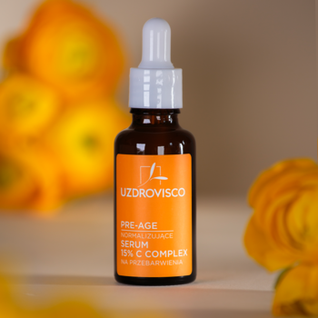 Uzdrovisco serum Pre-Age 15% C Complex w brązowej butelce z pipetą, na rozmytym tle kwiatów.