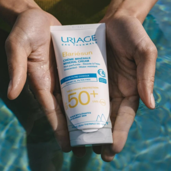 Zbliżenie na biały krem mineralny Uriage Bariésun SPF 50+ w tubce, trzymany dłońmi w wodzie.