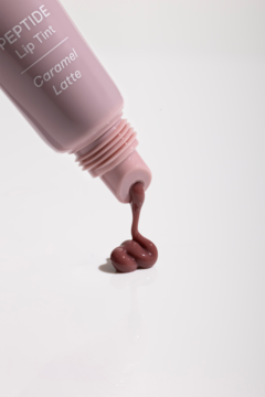 Różowa tubka Peptide Lip Tint Caramel Latte, z której wyciskany jest gęsty, brązowo-beżowy tint nawilżający na biel.