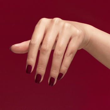 Dłoń z błyszczącymi, bordowymi paznokciami wzmocnionymi odżywką OPI Nail Envy Tough Luv na czerwonym tle.