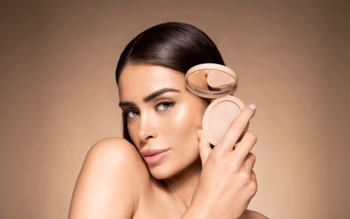 Kobieta prezentuje rozświetlacz Pupa Wonder Me Glow 101 Rose Gold w otwartej i zamkniętej puderniczce obok twarzy.