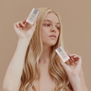Blondynka trzyma dwa małe, jasne opakowania Glov Magnet Cleanser do pędzli, na neutralnym tle.